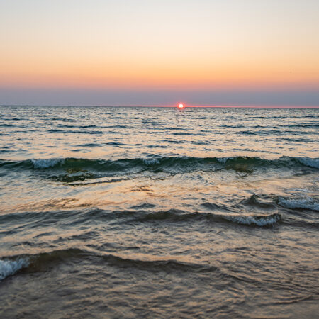 A view of the beauty of a sunset on Lake Michigan with the waves crashing into the shore near Muskegon, Michigan. The warm hues of the sky and water are reflected in the calm lake, creating a truly magical scene. 